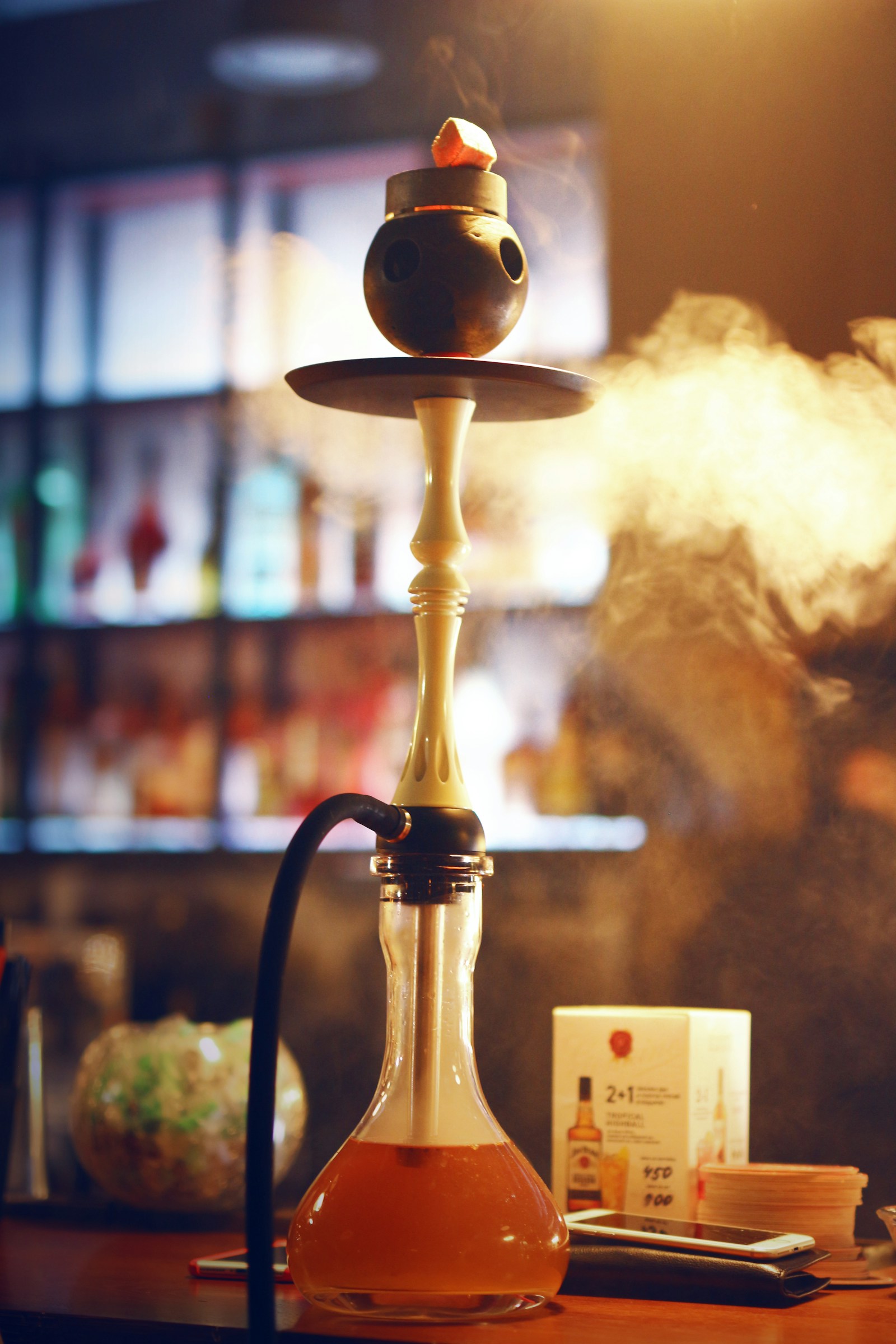 A hookah with a glowing coal and drifting smoke in front of a warm-lit bar
